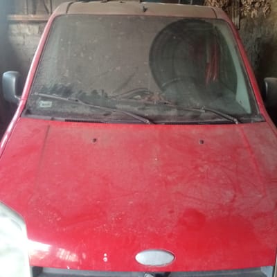 A red car, covered in dust, is parked inside a dimly lit garage or shed. The car's hood and windshield are visible from a frontal perspective. The windshield is heavily smudged with dirt, obscuring the view of the interior, though a large circular object, possibly a spare tire, is visible through it. A side mirror is attached to the driver's side of the car. The environment appears to be a storage area, possibly a rural setting, given the brick wall visible in the background. No people are present in the image. The location is identified as Slobidka, Ukraine.