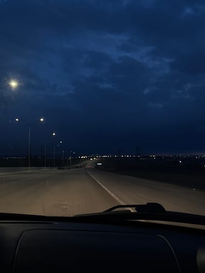 The image captures a nocturnal or late twilight scene from the perspective of a moving vehicle, looking towards the front passenger side. The scene is dominated by a dark blue sky with indistinct, shadowy clouds occupying the upper left quadrant of the frame.

A long, straight stretch of road, possibly a highway or bridge, runs horizontally across the middle and right side of the image, appearing to recede into the distance. The road surface is dimly lit, likely by streetlights and the vehicle's headlights, with reflections of light visible on its surface. Along the edge of the road, a series of streetlights with bright, circular glows extend into the distance, illuminating the immediate area. In the far distance, a faint line of numerous small, bright lights suggests a city, town, or industrial area along the horizon, creating a stark contrast against the dark sky and landmass.

Part of the vehicle's interior, specifically a dark dashboard or door panel, and a windshield wiper are visible on the far right edge of the frame, confirming the vantage point from inside a car or similar vehicle. No people are visible in the image.

The time of day is clearly night or very late dusk due to the deep blue sky and the reliance on artificial lighting. The presence of clouds indicates a non-clear sky, but no other specific weather conditions like rain or fog are apparent. There is no visible text in the image. Given the context of Bol'shaya Mos', Russia, this could be a view of a road or bridge leading towards or through a settlement.