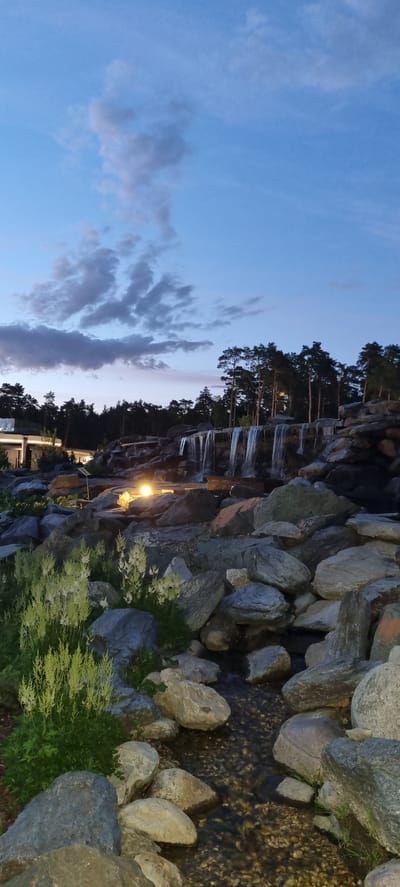 The image depicts a rocky waterfall scene with a stream flowing through it. The waterfall is constructed from large, grey rocks, and water cascades down in thin streams. Lush green vegetation, including grasses and small bushes, grows among the rocks. A single lamp post illuminates the area with a warm yellow glow, highlighting a portion of the rocky landscape and the waterfall. In the background, a dense line of evergreen trees, likely pines, forms a dark silhouette against the twilight sky. The sky itself is a gradient of blues, with scattered clouds that are tinged with pink and purple hues, indicating it is either dawn or dusk. The water in the stream is dark and appears shallow, with pebbles visible on the streambed. The overall impression is one of a tranquil, naturalistic setting, possibly a park or a landscaped garden, enhanced by artificial lighting. There are no people or animals visible in the image. The scene evokes a sense of peacefulness and natural beauty, with the warm light adding a touch of serenity to the cool tones of the sky and rocks. The image orientation is landscape, but the content appears rotated 90 degrees clockwise.
