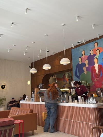 The image shows the interior of a café, possibly in Zamankul, Russia.
**People:** A barista stands behind a counter, facing right and attending to equipment. A customer with long, red hair stands at the counter, facing away from the camera. Further into the café, two seated patrons appear to be conversing.
**Subjects:** The focus is on the café's interior design.
**Objects:** A counter with coffee machines and supplies dominates the foreground. Hanging globe-shaped lights provide illumination. Artwork adorns the walls, including a large mural of figures in traditional dress above the counter, and a painting behind the customer. Seating consists of leather sofas.
**Actions:** The barista is likely preparing a beverage. The customer stands at the counter, possibly placing an order. Seated patrons are conversing.
**Interactions:** A transaction between the customer and barista is implied. The patrons are interacting in conversation.
**Environment:** The café features a modern, eclectic design. Wood paneling and a textured wall provide architectural details. Track lighting is visible on the ceiling. The flooring consists of brown and white stripes.
