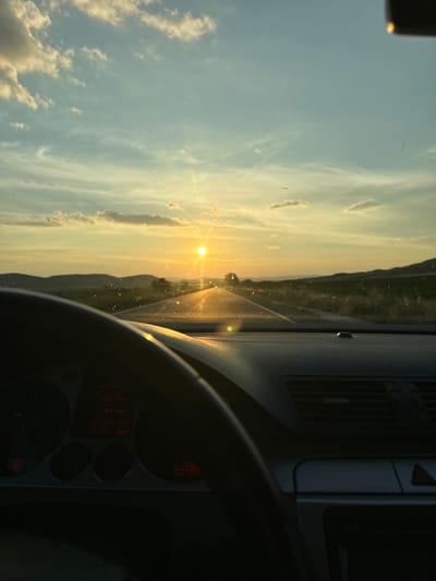 An interior view from the driver's seat of a car traveling on a road towards a sunset. The sun is a bright orb low on the horizon, casting a golden glow across the sky and reflecting off the asphalt road ahead. The sky is a gradient of blue and yellow, with scattered clouds. The landscape on either side of the road is hilly and appears to be rural, with some vegetation and distant trees. The dashboard of the car is visible in the foreground, with part of the steering wheel on the left and an air vent on the right. The car's instrument cluster displays red numerical readings, including "23565" likely indicating mileage. The image was captured on the road to Drugan, Bulgaria, likely during golden hour.
