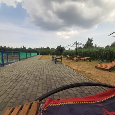 This is a wide-angle, eye-level shot of an outdoor recreational area. A paved walkway, made of gray interlocking stones, occupies the lower left and foreground of the image. To the left of the walkway is a body of water, bordered by a blue and green fence. Beyond the fence and water, lush green trees form a dense treeline.
The walkway leads to an area with sparse, dry grass and several amenities. There is a dark metal bench with a trash receptacle next to it. Further back, three wooden lounge chairs with black metal frames are arranged on the grass. Two large, black, umbrella-like structures, likely shade structures or canopies, are also present. Behind these structures, tall, reedy plants grow densely, bordering the water. In the distance, more trees are visible against a cloudy sky. The overall impression is of a somewhat unkempt but functional outdoor leisure space. The image does not contain any discernable human presence. The location is identified as Dubrivka, Ukraine.