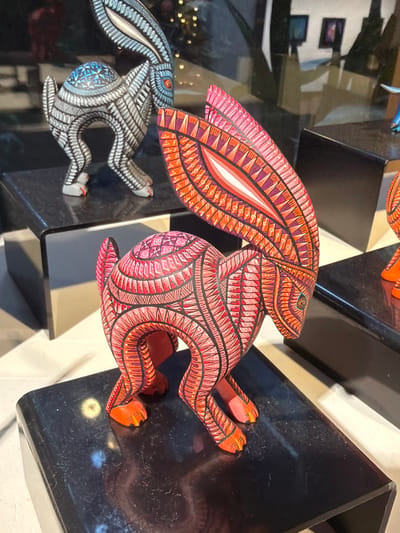 The image displays several vibrant and intricately patterned alebrijes, which are Mexican folk art sculptures, predominantly featuring rabbit figures. The central and largest subject is a red and orange rabbit, adorned with elaborate geometric and abstract designs in pink, purple, white, and black across its body, ears, and limbs. It is depicted in a dynamic, seemingly leaping pose.
Partially visible in the lower left is another smaller rabbit alebrije, colored in shades of light blue, grey, white, and black, also covered with complex patterns. Both sculptures are presented on glossy, black, reflective platforms arranged geometrically, which mirror parts of the figures and the surrounding environment. The background includes blurred reflections of an indoor space, possibly showing windows or architectural elements, along with small, circular lights.
The scene suggests an indoor setting, likely a gallery, artisan shop, or display area. The detailed craftsmanship, rich colors, and fantastical animal forms are characteristic of alebrijes, a traditional Oaxacan art form, consistent with the location context of San Martín Tilcajete, Mexico. The items are displayed for admiration or sale, with no human activity visible. The lighting is bright, highlighting the intricate details of the sculptures. No discernible text is present in the image.