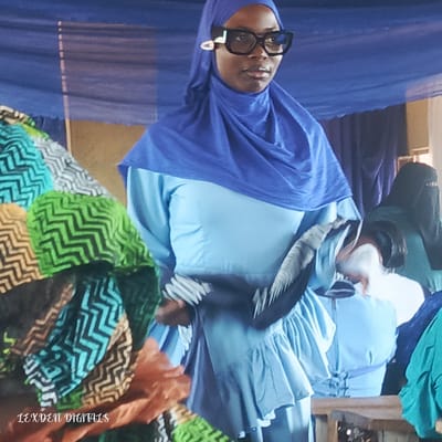 The image shows a group of people, primarily women, at an indoor event. One woman in the foreground, wearing a vibrant blue hijab and a light blue dress with ruffles, is the main subject. She has large, black, geometric-shaped glasses and is holding a patterned fan or accessory. Her posture suggests she is either posing or observing something. Other women are visible in the background, also wearing hijabs and modest attire in various shades of blue and green.

The setting appears to be a gathering or celebration, likely in Nigeria given the location context. The backdrop consists of draped blue fabric, creating a festive atmosphere. The lighting is somewhat dim, suggesting an indoor event possibly taking place during the day or evening. The overall mood seems to be one of quiet observation and perhaps a fashion show or cultural presentation, given the stylish attire and accessories.

The fabric in the foreground, to the left of the main subject, features a striking pattern of green, black, and beige zigzags. Visible text at the bottom left corner reads "LEXDEN DIGITALS". The focus is on the people and their clothing, highlighting cultural fashion and personal style within the context of a community event.