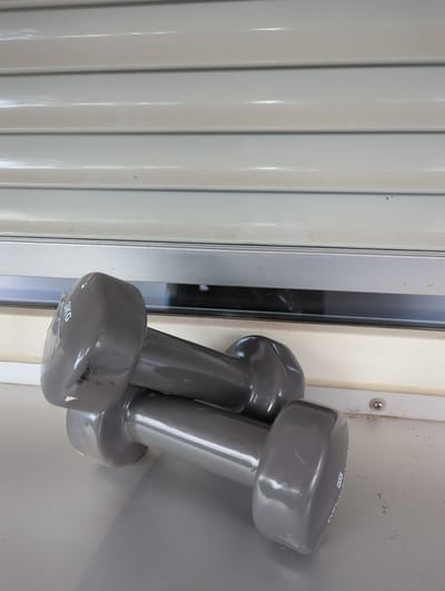 Two grey dumbbells are leaning against a light-colored vertical surface. The dumbbells appear to be of similar size and weight, with one slightly in front of the other. The one in the foreground has "3KG" printed on its end in white text. The background consists of a ribbed, light-colored surface, possibly a wall or a door, and a vertical pane of glass or plastic. The lighting suggests an indoor setting, possibly during daylight hours. There is a screw visible on the vertical surface to the left of the dumbbells. The overall scene is simple and focuses on the exercise equipment. There are no people or any indication of an activity taking place, other than the presence of the dumbbells themselves.