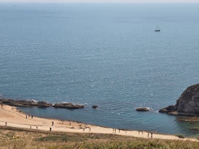 This aerial view captures a sun-drenched coastal scene at Lulworth Cove, West Lulworth, United Kingdom. The expansive, choppy blue sea fills the upper two-thirds of the frame, with a distant sailboat visible on the horizon. The lower portion of the image reveals a sandy beach nestled against dramatic, rocky cliffs.
Several individuals are scattered across the beach and a well-trodden dirt path that runs parallel to the shoreline. They appear to be leisurely walking and exploring the area, with no discernible interactions between them. The beach itself is sparsely populated, with only a few small groups or individuals visible. The surrounding landscape is arid and grassy, with dry, yellowish vegetation in the foreground. The cliffs are rugged and appear to be composed of sedimentary rock, with some darker, greener seaweed visible in the shallow, clear water near the shore. The overall impression is one of a popular tourist destination on a clear, sunny day.