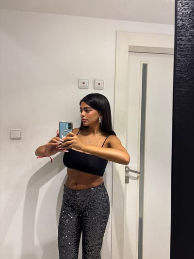 The image shows a young woman standing in a bathroom and taking a selfie. She is wearing a black crop top and black sequined pants. The woman's dark hair is pulled back, and she is wearing hoop earrings. She is holding her phone in her right hand and looking at the camera. There are two light switches on the white wall to the left. On the right is a white door with a silver handle. Behind her, a mirror reflects the scene. The coordinates 31.916, 34.867 are in the city of Ramla, Israel, which is 35.1 km south-east of Tel Aviv.