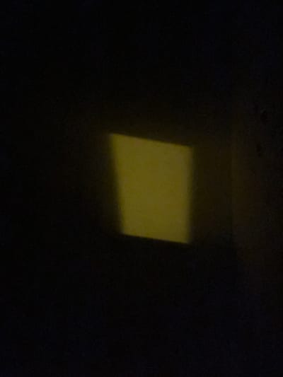 A square of yellow light is projected onto a dark surface, possibly a wall. The light source and the surface are not visible. The image is dark and grainy, with the yellow square being the only discernible feature. There is no indication of location or context beyond what is visually present.