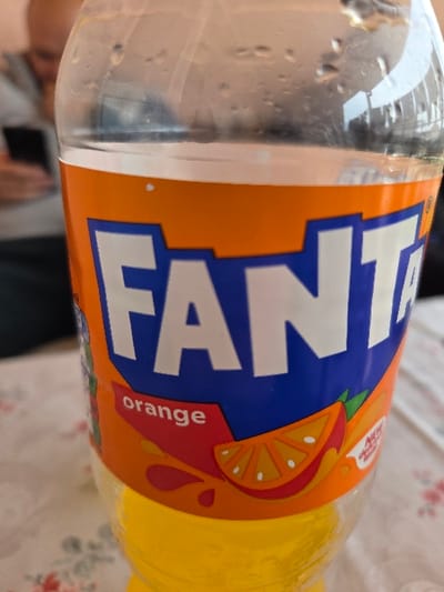 A clear plastic bottle of Fanta Orange soda, partially full with a yellow-orange liquid and exhibiting external condensation droplets, is positioned centrally on a light-colored surface covered by a floral patterned tablecloth in Negorci, North Macedonia. The bottle's label displays the "FANTA" logo in white on a blue background, with the word "orange" and graphic representations of orange slices and juice splashes on an orange field. In the blurred background to the upper left, a person with short hair or a bald head, wearing a light-colored top, is visible looking down and engaging with a dark-colored smartphone held in their hands.