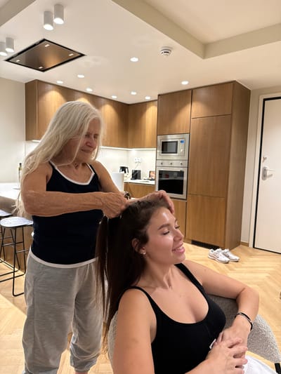 Here is a description of the image:
The image shows two women indoors, one older and one younger. The older woman, with long white hair, is standing and appears to be styling the younger woman's hair. The younger woman is lying down, facing the older woman. Both are wearing casual clothing.
The location appears to be a modern kitchen or living space, identifiable by wooden cabinets, built-in appliances, and contemporary lighting fixtures. There is no specific information to determine if the image was taken in the Municipal Unit of Elliniko, Greece.