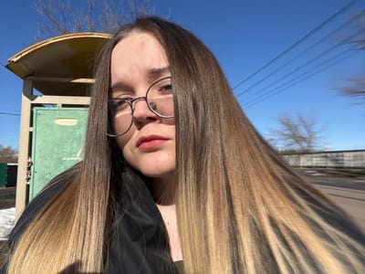 A young woman with long, straight, dark brown to blonde ombre hair, wearing round metal-frame glasses and dark clothing, is centered in a selfie, looking directly at the camera. Her lips are adorned with red lipstick, and a small blemish is visible on her forehead. To her left, partially obscured, is a weathered structure featuring a yellow, curved corrugated metal roof atop a rectangular, green-painted panel, resembling a bus stop or small shelter. A white paper is affixed to the green panel. Bare tree branches are visible above this structure against a clear blue sky. Multiple utility lines are discernible in the upper right quadrant. Patches of snow lie on the ground in the lower left, indicating cold conditions. In the blurred background to the right, additional bare trees, a grey structure consistent with a fence or building, and a road or dirt path are present. This outdoor scene is located in the city of Большевик, Russia.