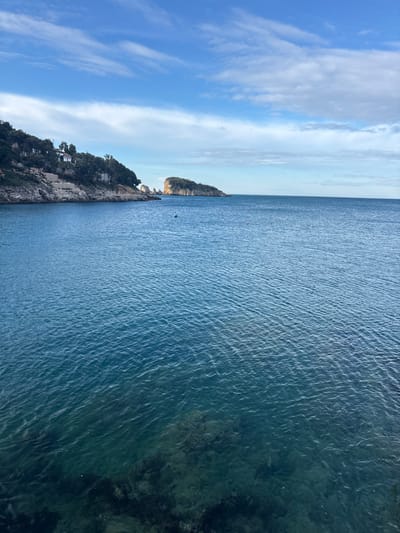 The image depicts a coastal scene in Kruče - Kërruçi, Montenegro, viewed from an elevated perspective overlooking the sea. The majority of the frame is dominated by a body of deep blue-green water, exhibiting minor surface ripples. The water in the immediate foreground is clear enough to reveal submerged rocky formations and darker patches of what appears to be marine vegetation on the seabed.
The left mid-ground features a rugged, rocky coastline densely covered with green trees and shrubs. Several white, multi-story buildings are observable nestled into the hillside among the vegetation, with some terraced structures or retaining walls visible below them. Extending from this coastline, towards the center-right, are smaller, jagged rock formations protruding from the water. Further out in the mid-ground, a larger, prominent rocky island or peninsula, also covered in green foliage, is visible.
Two buoys are present in the water: a green conical buoy in the left mid-distance and a smaller, orange-red cylindrical buoy positioned further right. The horizon line separates the water from a light blue sky, which contains scattered white, wispy stratocumulus clouds. No individuals, vessels, or direct actions are depicted.