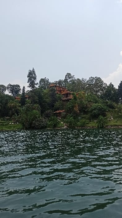 The image displays a panoramic view of a lush, forested hillside rising from the edge of a large body of water under an overcast sky in Bwishyura, Rwanda. The foreground consists of dark, rippled water, exhibiting subtle reflections of the sky and land. On the hillside, multiple structures with reddish-brown roofs and visible wooden elements are integrated into the dense green foliage. These buildings appear to be multi-story and tiered, possibly lodges or residences, with some featuring visible balconies or verandas. A smaller, single-story structure with a similar roof is situated closer to the water's edge on the lower left side of the hill. The vegetation is abundant, featuring various trees and shrubs. No individuals, explicit actions, or direct interactions are discernible within the frame. The sky is largely pale grey, with some brighter white cloud formations visible towards the upper right.