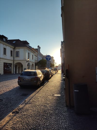 The image captures a street scene in Kolín, Czechia, bathed in the warm, low light of the setting sun. The cobblestone street is lined with parked cars, the closest being a beige station wagon with a Czech license plate. The sun casts long shadows across the uneven pavement, highlighting the textured surface. On the left, a row of historic buildings with archways and ornate windows stands tall, their facades reflecting the golden hour glow. A lamppost is visible in front of one of the buildings. A blue traffic sign with a bicycle symbol and arrows indicating turns is also present. The right side of the image is dominated by the side of a building, casting a large shadow that stretches across the street. Further down the street, more cars are parked and the sun is partially obscured by a distant building. The sky is a clear, light blue.