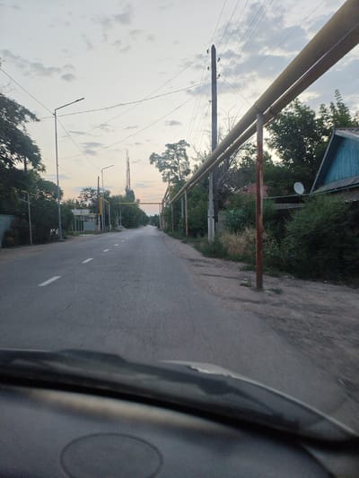 The image shows a street scene in the city of Shu, Kazakhstan, under a cloudy sky at dusk. The perspective is from inside a vehicle, looking forward through the windshield. The road is paved with asphalt and has dashed white lane markings. On either side of the road are trees, bushes, and some residential structures. A prominent feature is a yellow pipeline supported by metal posts, running parallel to the road. Utility poles with overhead wires are also visible. In the distance, the road appears to curve slightly. The overall impression is of a rural or semi-urban street in a temperate climate.