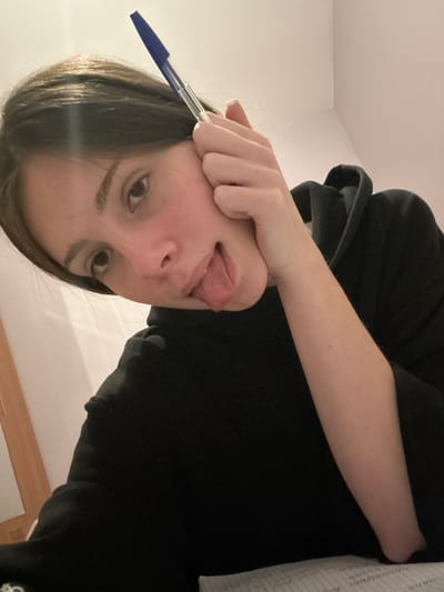 An upside-down selfie of a person with dark hair sticking their tongue out. They are holding a blue pen to their face. The person is wearing a black hooded sweatshirt and their arm is visible. A wall and a wooden panel are visible in the background. A notebook is partially visible in the upper right corner.
