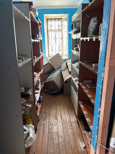 This media file shows a cluttered room with multiple shelves filled with various items. The shelves are wooden, with some appearing worn and others painted white. Many of the shelves have labels with numbers, such as 56, 60, 61, 64, 65, 66, 67, 68, 71, 72, 73, 74, and 21, 22. These numbered labels suggest the shelves might be part of a postal sorting facility, a storage system, or mailboxes.
The shelves are filled with a variety of objects, including cardboard boxes of different sizes, plastic bags containing items, and what appear to be rolls of paper or posters at the top of the shelves. Some shelves contain smaller, more organized items, while others are overflowing with disorganized clutter. In the foreground, there are also several large cardboard boxes piled up, some of which are open.
A window with a decorative metal grate is visible in the background, letting in natural light. The walls appear to be painted blue, and the floor is made of old wooden planks that show signs of wear and tear.
There are no people visible in the image. The scene appears to be indoors, possibly in a dimly lit area due to the presence of natural light from the window. The overall impression is one of disarray and accumulated items, suggesting a place that is either actively in use and being filled, or a storage area that is neglected. The location is identified as Vileyka, Belarus.