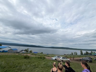 The image captures an outdoor scene at the edge of a body of water, likely a lake or reservoir, with a shoreline in the distance. The sky is overcast with numerous clouds. In the foreground, a grassy embankment slopes down towards the water. Several people are visible in the immediate foreground, some facing away from the camera.
Further back, near the water's edge, there are several cars parked on a dirt area. Adjacent to the parking area are buildings with blue roofs, suggesting a recreational area or a small settlement. One building is larger and appears to be a structure with windows, while a smaller, shed-like building is next to it. A blue tarp structure is also visible on the left side of the image.
On the water, a house-like boat or barge is floating, and a smaller boat is also present. In the middle ground and background, the opposite shoreline features a treeline and rolling hills.
The location is identified as Sergeevka, Russia. The scene appears to be a waterfront area, possibly a park, a recreation spot, or a small village on the water. The overall atmosphere is one of a cloudy, possibly cool day.
