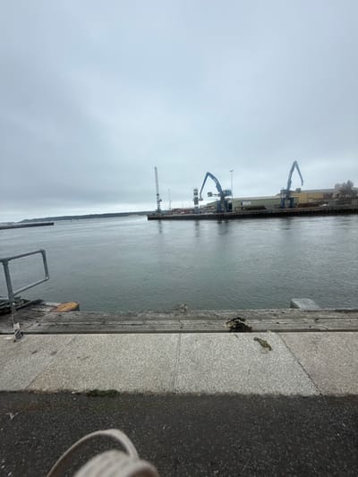 The image depicts a view from a pier at the Port of Poole in the United Kingdom. The foreground shows a wooden pier with a metal railing on the left, leading to a paved area. Beyond the pier is a wide expanse of water, likely Poole Harbour, with a distant shoreline covered in trees. On the far side of the water, industrial structures are visible, including tall cranes, warehouses, and shipping containers, characteristic of a working port. The sky is overcast and grey, suggesting a cool, possibly damp, day. There are no people visible in the scene.