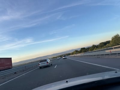 The image captures a view from a vehicle traveling on a highway, likely near Malo Buchino, Bulgaria. The sky is a clear, pale blue with wispy clouds and a faint contrail from an aircraft. The highway is paved and has white lane markings. Several cars are visible on the road, including a silver station wagon in the foreground with license plate "CB 9220 BH". A red truck is parked on the left side of the highway. The highway is bordered by a metal guardrail, and beyond that, there is a landscape with rolling hills and trees. The lighting suggests it is either early morning or late afternoon.