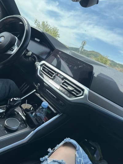 The image provides an interior view of a modern car, specifically a BMW due to the visible steering wheel logo and dashboard elements, taken from the passenger's perspective. The legs of a person wearing distressed denim shorts are prominent in the foreground on the passenger side.
Through the windshield, a bright blue sky with scattered white clouds is visible, along with distant green hills or mountains, indicating clear and sunny daytime weather. A street light or sign pole can also be seen outside. The digital instrument cluster displays "D7 +16.5" and "76."
The central dashboard features a large infotainment screen showing "11:45" as the time, along with "Music collection," "Previous track," and "Next track." Below it are silver-accented air vents and climate control buttons, including "MAX" and "A/C." The center console between the seats holds a "VIVA" brand water bottle, a "GoPro" camera, a white disposable cup, and a charging cable. The gear selector area displays "P," "R," and "OFF," while the iDrive controller is surrounded by buttons labeled "MEDIA," "HOME," "MAP," "NAV," "OPTION," and "BACK." The car's interior trim includes a textured, possibly carbon-fiber-like pattern on the dashboard and console. The scene suggests a casual journey or stop in Madrets, Bulgaria, with the vehicle either parked or slowly moving.