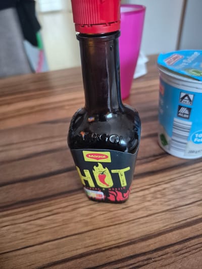 The image shows a close-up of a bottle of Maggi Hot sauce and a container of Aldi dairy product on a wooden table. The bottle of Maggi Hot sauce is dark brown glass with a red cap and a black label featuring the word "HOT" in large yellow lettering with a red chili pepper design. The Aldi container is white with a blue lid and features the Aldi logo. In the background, a pink plastic cup is visible, and a blurry figure wearing black and green clothing is present. The scene is indoors, and the lighting suggests it is daytime. There is no specific indication of the location in Barbing, Germany.