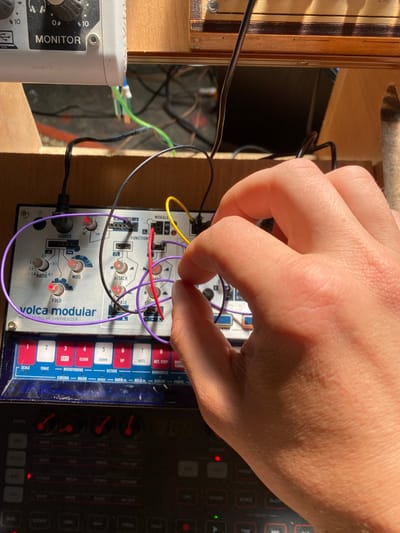 The image shows a close-up of a person's hand interacting with a "volca modular" synthesizer. The synthesizer is silver with various knobs and patch points, some of which are connected with colored patch cables (purple, red, and yellow). The hand is holding a purple patch cable, appearing to insert or remove it from a jack on the synthesizer. The synthesizer is part of a larger setup, with other electronic music equipment visible in the background, including a white unit labeled "MONITOR" and what appears to be a wooden rack. The lighting suggests an indoor setting, possibly a home studio. There is no indication of the location being in Лугаваслабадскі сельскі Савет, Belarus.