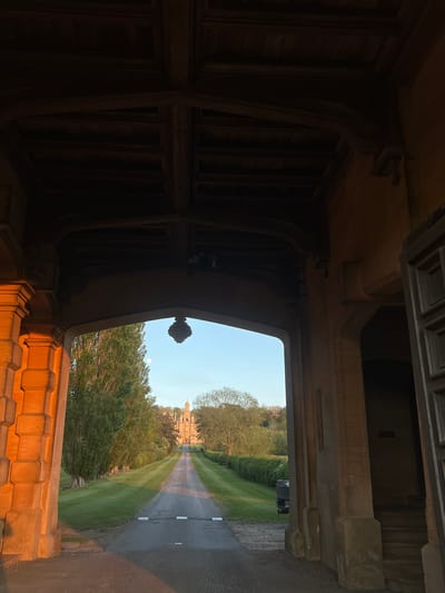 The media file presents a view from within a dark, ornately structured archway or tunnel, looking out onto a bright, expansive landscape. The image is initially oriented vertically, appearing rotated 90 degrees counter-clockwise.
**Subjects:**
The foreground is dominated by the interior of the archway, featuring dark wooden beams, panels, and intricate carvings along the ceiling and walls. Stone elements with detailed moulding and a decorative finial are also visible. In the midground, seen through the archway's opening, is a long, straight tarmac driveway, flanked by meticulously maintained green lawns and various trees. In the far distance, at the end of the driveway, a grand, multi-turreted or towered building, likely a stately home or castle, stands prominently. The sky above is a clear, pale blue, with a faint, small moon visible in the upper right. Part of a stone balustrade or wall in the immediate right foreground is illuminated by warm, golden light. A dark-colored vehicle (possibly an SUV) can be faintly discerned on the driveway further down.
**Activity or event taking place:**
No specific activity or event is depicted; the scene captures a static architectural and landscape view.
**Notable details:**
The dramatic contrast between the shadowy interior of the archway and the brightly lit exterior is a key feature. The golden illumination on the stonework in the immediate foreground, combined with the pale sky and visible moon, suggests either early morning or late afternoon/early evening (golden hour). The clear sky indicates fair weather. The architecture of the archway and the distant building, along with the manicured grounds, strongly suggest a historic country estate or grand residence. Given the location context of South Kesteven, United Kingdom, this likely depicts a significant historical property in the region. The overall atmosphere conveys grandeur, history, and a sense of peaceful observation.
**Visible text:**
There is no visible text in the image.