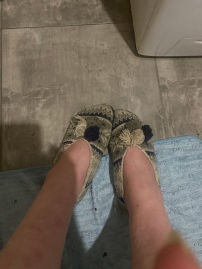 A close-up, top-down shot shows a pair of feet wearing patterned slippers. The slippers are a knit material with a cream, blue, and dark gray geometric pattern, and each has a small pom-pom accent near the ankle opening. The feet are resting on a textured blue bath mat, which is placed on a gray tiled floor. The background includes a sliver of a white appliance, possibly a washing machine or dryer, and a shadow cast from the left. The lighting is dim, creating some shadows on the floor and around the feet. The scene is indoors, likely in a bathroom or laundry area.