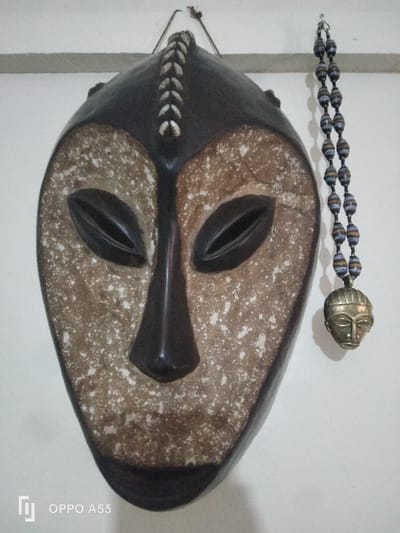 The image displays a dark brown and beige wooden mask, vertically suspended against a plain, light-colored wall in Riabai ward, Kenya. The mask features a prominent dark brown vertical strip down its center, adorned with a series of small, white, ovular embellishments. Its eye sockets are elongated and dark brown, while the nose is a prominent, angular dark brown. The wider facial planes are a lighter beige, mottled with white speckles. To the right of the mask, a double-strand necklace hangs from the wall. This necklace is composed of numerous small, cylindrical beads, exhibiting various tones of blue, beige, brown, and white, consistent with rolled paper construction. Attached to the bottom of the necklace is a small, metallic pendant shaped like a human head, featuring stylized facial features and a brass-like patina. Both items are affixed to the wall via unseen hooks or fasteners. The environment is an interior space with diffuse lighting.