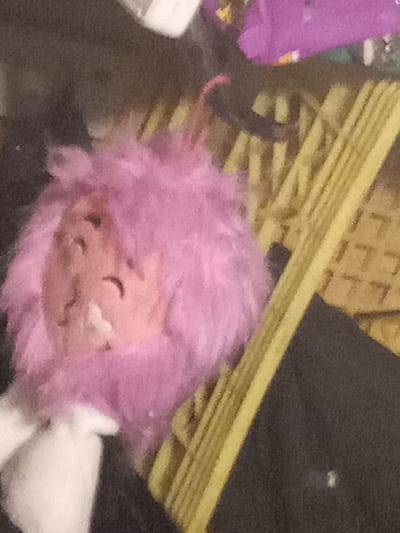 A pink fluffy toy with a white face is laying on a woven surface made of many thin strips of yellow material. The toy is facing right, and its right eye is visible. The toy is partially obscured by a black surface. This scene is most likely located in a private residence in Kitchener, Canada, based on the provided coordinates.