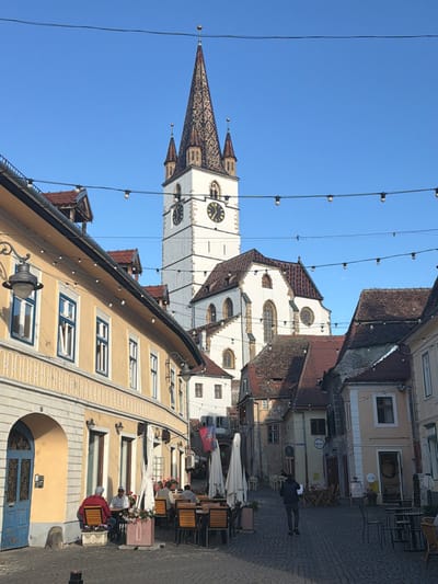 The image depicts the Evangelische Stadtpfarrkirche, a prominent landmark in Sibiu, Romania. The church's tower, with its clock faces and patterned roof, rises against a clear blue sky. Strings of lights crisscross the foreground. Below, there are buildings lining a cobblestone street, one with yellow facade and blue window frames, others with red roofs. Tables and chairs of an outdoor cafe are set up, occupied by people, some wearing hats. A person in a dark jacket walks along the street. A flag flies from one of the buildings.