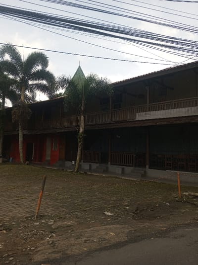 The image displays a two-story building in Chori, Nigeria, under an overcast sky. The structure features a brown tiled roof, from which a green, conical spire rises prominently near the center. The upper floor is characterized by an open-air balcony extending the building's length, enclosed by dark wooden railings. The ground floor's facade presents distinct sections: the left side consists of two segments painted red, each with a door and a window. The right side exhibits darker walls, possibly dark blue or green, with similar wooden railings, behind which interior seating arrangements are visible. Multiple tall palm trees are situated in front of and around the building. The ground in the foreground transitions from discolored paving stones and exposed dirt patches near the building to an unpaved dirt area, culminating in a paved road on the lower right. Two wooden stakes with orange tips, connected by a thin chain, are positioned in the immediate foreground. Numerous black utility wires are suspended across the upper portion of the frame against the sky. No individuals are present in the scene.