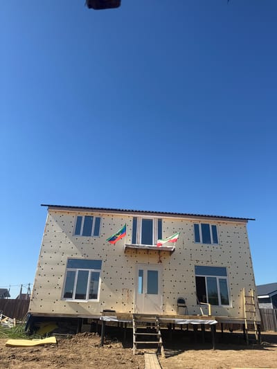 A two-story, unfinished house is shown under a clear blue sky in the city of Matchino, Russia. The house is covered in a light-colored insulating material with black circular fasteners. Two flags, one with green, white, and red stripes, and another with green, white, red, and blue stripes, are attached to the front of the house. The house has several windows and a white door with a small overhang. A wooden deck with stairs leads up to the entrance, and a ladder leans against the side of the house. Construction materials, including yellow foam insulation, are visible on the ground. A chair and a piece of wood are on the deck. The surrounding area is a dirt construction site with a wooden fence in the background.
