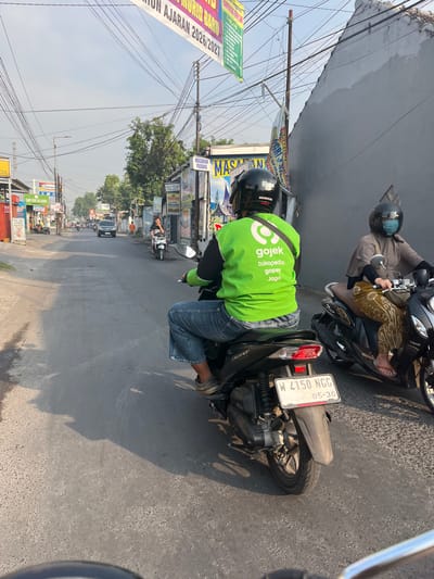 A side view of two Gojek motorcycle couriers riding down a paved street in Kedungkendo, Indonesia. The courier in the foreground is wearing a bright green Gojek jacket and blue jeans, while the courier behind them is wearing a grey headscarf and patterned pants. Both are wearing helmets. The street is lined with buildings and signs, and power lines crisscross overhead. The overall scene depicts a typical day in an Indonesian urban environment.