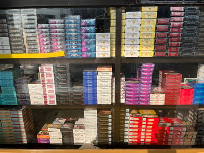 The image shows a storefront display of various cigarette brands. The storefront appears to be a small retail shop, possibly a kiosk or a corner store, located in the city of Bulu, Indonesia. The cigarettes are neatly stacked on shelves behind glass. Brands visible include Djarum, Camel, Magnum, Esse, Lucky Strike, Diplomat, Surya Pro, Marlboro, and Rock Star, among others. The display is well-organized with different brands grouped together. The lighting suggests an indoor setting, with reflections on the glass obscuring some details. The overall scene depicts a typical retail environment for tobacco products in Indonesia.