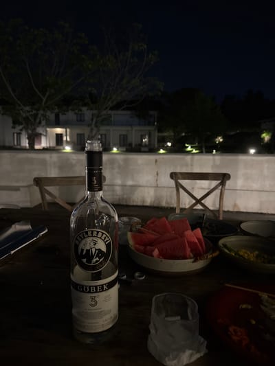 The image depicts a table setting at night. A bottle of Beylerbeyi Göbek rakı lies on the table amidst plates of food, a bowl of sliced watermelon, a glass with ice, and miscellaneous objects. In the background, lights suggest a building or street scene at night. The location is not identifiable as Jinhua, China, from the information provided.