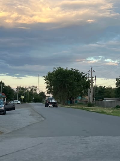 The image depicts a roadside scene in Zimovniki, Russia, likely during the late afternoon or early evening, as indicated by the soft, diffused light illuminating the sky. The sky is a dramatic canvas of clouds, with streaks of golden light breaking through the predominantly gray and blue tones, suggesting a transition from day to night or a clearing after rain.
The foreground shows a gray asphalt road curving into the distance. On the right side of the road, there is a concrete embankment. A few vehicles are parked along the side of the road. One is a white car, and behind it, a dark object, possibly a dumpster or a large piece of equipment, is visible. Further down the road, a black and white truck is parked.
On the left side of the road, a line of utility poles with wires stretches into the distance, a common sight in rural or suburban areas. Lush green trees and bushes line the road, creating a natural barrier between the road and what appears to be a building or structure partially visible in the background on the far left. The trees are dense and full, suggesting a temperate climate and a time of year with active vegetation growth. A yellow pedestrian crossing sign is also visible on a utility pole.
The overall atmosphere is one of quietude and everyday life, with no people overtly visible in the scene. The natural elements, particularly the sky and the vegetation, are prominent, creating a sense of place that is both tranquil and slightly dramatic due to the cloud formations. The image is oriented vertically, giving a strong sense of perspective along the road and up towards the sky.