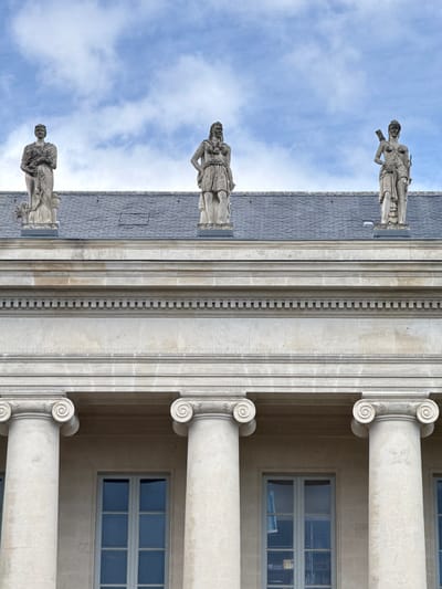 The image shows the exterior of a building with classical architecture. Three stone statues of robed figures stand atop the building's roof. The building features large columns with scrolled capitals and rectangular windows. The sky behind the building is blue with scattered clouds. This is the facade of the Nantes Museum of Fine Arts, located at Place du Lieutenant Colonel Sébert, 44000 Nantes, France.
