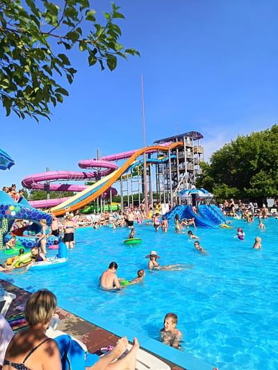 The image depicts a crowded water park on a sunny day, with a large blue swimming pool in the foreground. Several water slides are visible, including large purple spiral slides and a prominent orange and blue slide. People of various ages are enjoying the water, with some swimming, others sitting on inflatable toys, and children playing. There are also lounge chairs and umbrellas around the pool. Trees and a clear blue sky form the background. The location is specified as the water park in Yarovoye, Russia.