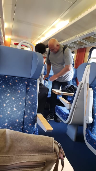 An interior shot of a train carriage in Margecany, Slovakia captures a bald, fair-skinned man, appearing to be in his late 50s or 60s, wearing a grey t-shirt and dark pants. He is standing and looking down, with his right hand on the handle of a black rolling suitcase. Another person, with dark, curly hair, is partially visible behind him. The train seats are upholstered in a blue fabric with a repeating pattern of orange and black geometric shapes. A small fold-down table is extended in front of the man, with a dark fabric item resting on it. A tan canvas bag is prominently displayed in the foreground, partially obscuring the seats. The lighting in the carriage appears to be overhead fluorescent fixtures, with orange accents on the ceiling and walls. Red curtains are visible in the background. The overall scene suggests a travel environment on a train.