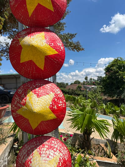 In Ia Hru, Vietnam, a vertical composition showcases four conical hats, reminiscent of traditional Vietnamese *nón lá*, painted red with yellow stars, stacked along a metal pole against a bright blue sky with scattered clouds. The hats are weathered, with red paint peeling to reveal a lighter underlayer. In the background, lush green foliage, including palm trees, borders a pale blue structure, possibly a pool. Further in the distance, houses with terracotta roofs are visible amidst more greenery. A black car is parked to the left, partially obscured by a planter. String lights with visible bulbs are suspended across the sky in the mid-ground. The scene captures an outdoor setting on a clear day.
