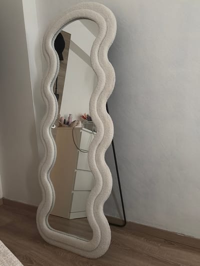 The image shows a fluffy, cloud-shaped mirror with a light-colored border. The mirror is positioned on a wall, reflecting a room interior. In the reflection, a white dresser with drawers is visible, along with some decorative items on top, including what appears to be a pink bottle and some flowers. A black object, possibly a bag or a piece of clothing, is also reflected in the mirror, placed on a shelf below it. The background of the room appears to be a wall with a subtle texture and a wooden floor. The lighting suggests it's daytime, possibly indoors with artificial light. The overall scene appears to be a bedroom or a dressing area. There is no visible text in the image.