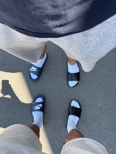 The image shows two pairs of legs and feet wearing white socks and Nike slide sandals on an asphalt surface. One pair of legs is wearing gray sweatpants with a dark blue stripe, and the other is wearing white shorts. One pair of sandals is black with a black Nike logo, and the other is navy blue with a white Nike logo. The photograph is taken from an overhead perspective. The image does not provide information to confirm a location in Kalotina, Bulgaria.