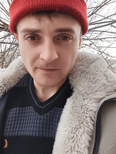 A close-up portrait features a male individual, appearing to be in his 20s to early 30s, looking directly into the camera. He has fair skin, brown eyes, and a light stubble on his chin and upper lip. His short brown hair is partially visible beneath a bright red knit beanie. He is dressed in a black V-neck sweater with horizontal stripes, alternating between solid black and dark grey waffle-knit patterns, and a small gold laurel wreath emblem is visible on the lower left chest. Over the sweater, he wears a light beige-tan jacket or coat featuring a prominent, thick light grey faux fur collar lining and a visible black zipper pull on the right side. The background is out of focus, revealing a dense network of bare tree branches against an overcast sky, suggesting an outdoor environment in winter or early spring. The scene is located in the city of Abay, Kazakhstan.