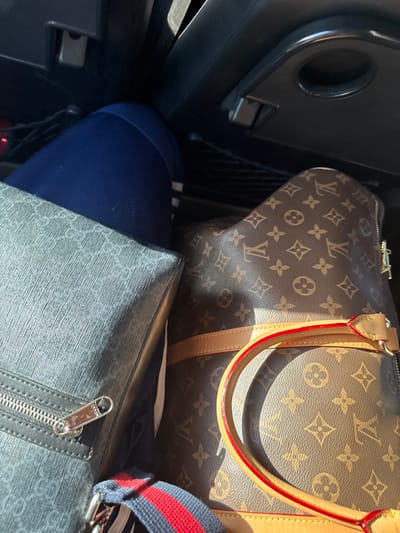 The scene depicts the interior of a vehicle during daylight, focusing on the contents of the rear passenger or cargo area. A person's upper left thigh, clothed in dark blue athletic pants with a white stripe near the knee, is visible in the upper left. Adjacent to the leg, on the left, is a rectangular gray-black bag featuring a patterned texture and a silver-toned zipper pull. To the right of the person's leg and the gray-black bag is a large brown travel bag with a distinctive pattern of interlocking "LV" monograms and floral motifs, characteristic of Louis Vuitton. This bag has light tan leather trim and handles with bright red edges. A strap with red and blue stripes is partially visible at the bottom center, possibly belonging to one of the bags or another item. The vehicle's interior features dark upholstery or plastic, with an overhead handle and a dark mesh pocket visible in the background. Overhead, a black fold-down tray with a circular indentation, possibly a cup holder, is partially visible. Natural light illuminates the objects, casting distinct shadows. No specific identifying markers for Pokrovskoye, Russia are visually present within the image; the environment is confined to the vehicle interior.