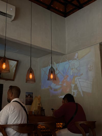 An indoor scene features three hanging lamps with caged Edison bulbs, descending vertically. Two people are partially visible at a table, one in a white shirt and the other in a red shirt, seemingly focused on their phones. A Spongebob Squarepants cartoon is projected onto the ceiling. A mirror and a small picture frame adorn the wall, alongside an air conditioner.