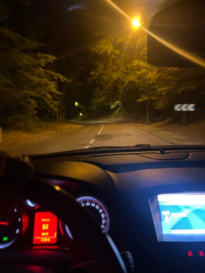 The image depicts a first-person view from inside a car at night, driving on a road lined with trees. The dashboard of the car is visible, showing a speedometer displaying "Vitesse 50 km/h" and a mileage of "224016". There is also a central infotainment screen displaying a navigation interface with the word "SÉLECTION" visible. The road ahead is illuminated by a streetlamp, and trees are present on both sides of the road, with some foliage illuminated by the headlights and streetlights. A blue and white chevron road sign is visible in the distance on the right side of the road. The overall impression is of a nocturnal drive through a semi-urban or rural area. The image is slightly blurry due to motion. The location is stated as the city of Maromme, France.