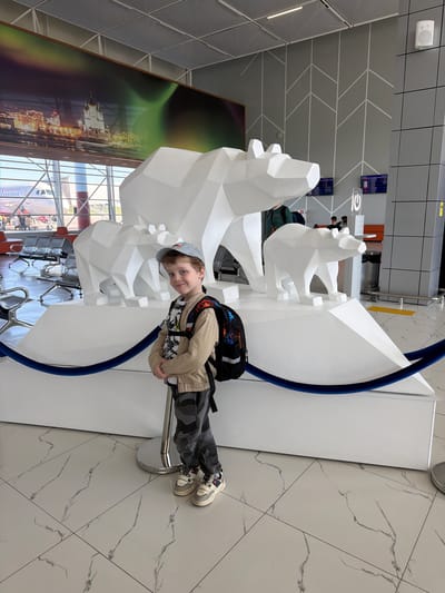The image depicts a young boy with curly hair, wearing a light tan jacket, a black and white patterned shirt, gray camouflage pants, a gray cap, and a backpack. He is standing beside a modern, abstract sculpture of white polar bears. The sculpture is part of an installation with a blue accent and is positioned in front of a large window and an artistic mural depicting Arkhangel'sk, Russia. The interior space appears to be a modern airport or transportation hub, evident from the marble-like flooring, clean lines, and seating arrangements in the background. The location is likely the Talagi Airport in Arkhangel'sk, Russia, indicated by the mural depicting the city.