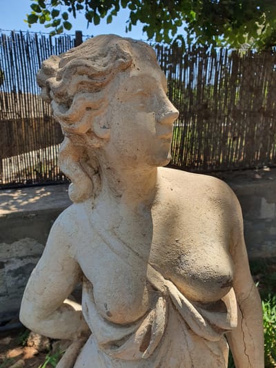 A weathered, light-colored stone statue of a semi-nude woman stands outdoors against a background of a rustic bamboo fence and lush green foliage. The statue is positioned in a garden setting, with sunlight casting shadows on its surface. The woman's hair is styled in curls, and she is draped in a piece of cloth that partially covers her body. In the background, a wooden fence made of vertical bamboo poles separates the garden from a shadowy area. The statue appears to be located in the municipal unit of Malia, Greece, likely in a private garden or courtyard given the outdoor setting and decorative statue.
